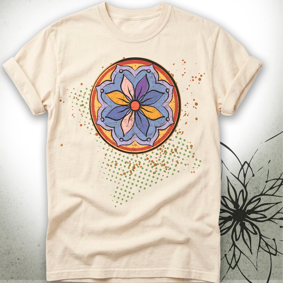 A light beige crewneck t-shirt featuring a colorful circular mandala graphic on the chest, displayed against a textured gray background with a black floral sketch on the right.