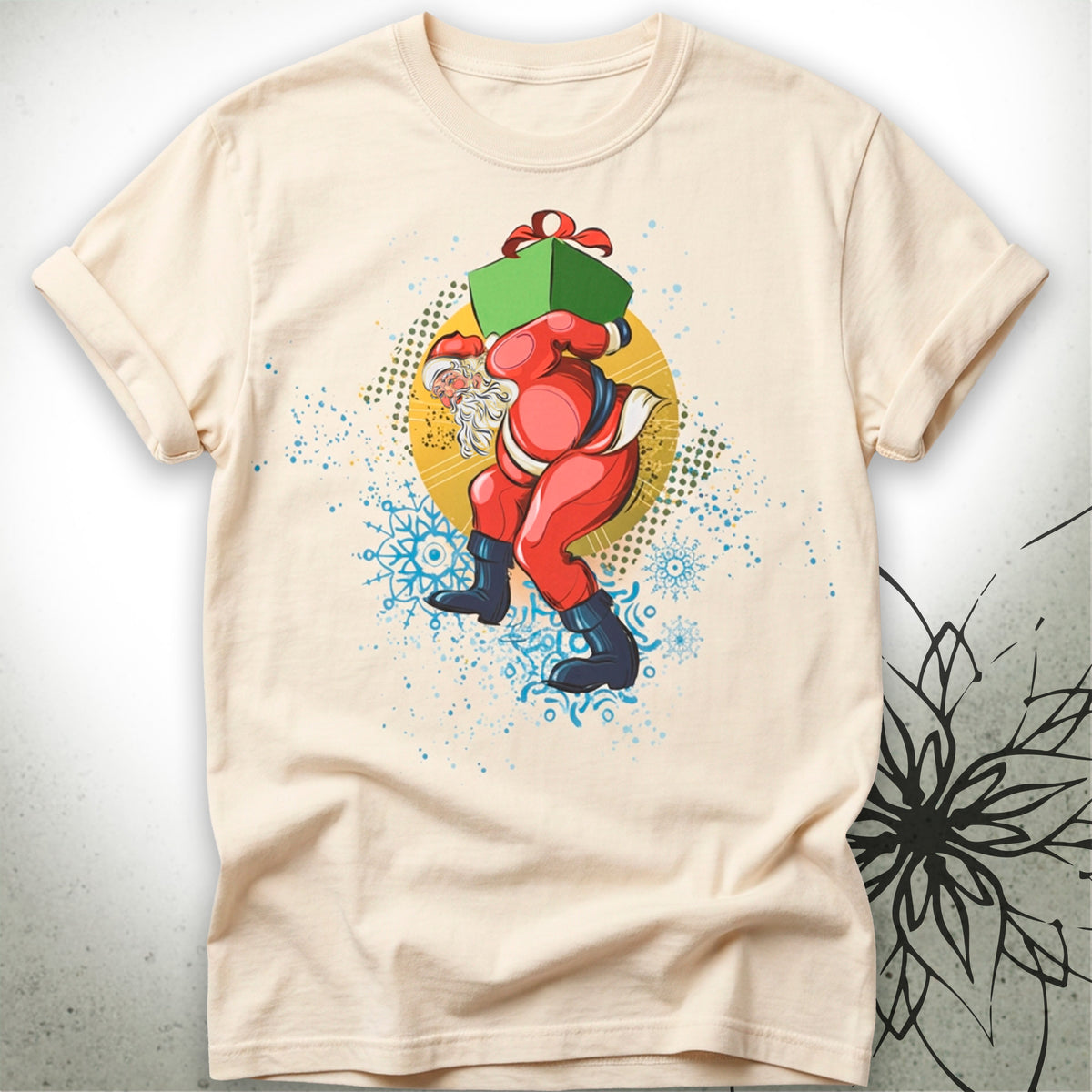 A light beige crewneck t-shirt featuring a colorful graphic of Santa Claus carrying a green gift box on his shoulders, set against a white background with a subtle gray textured pattern and black floral design on the right.