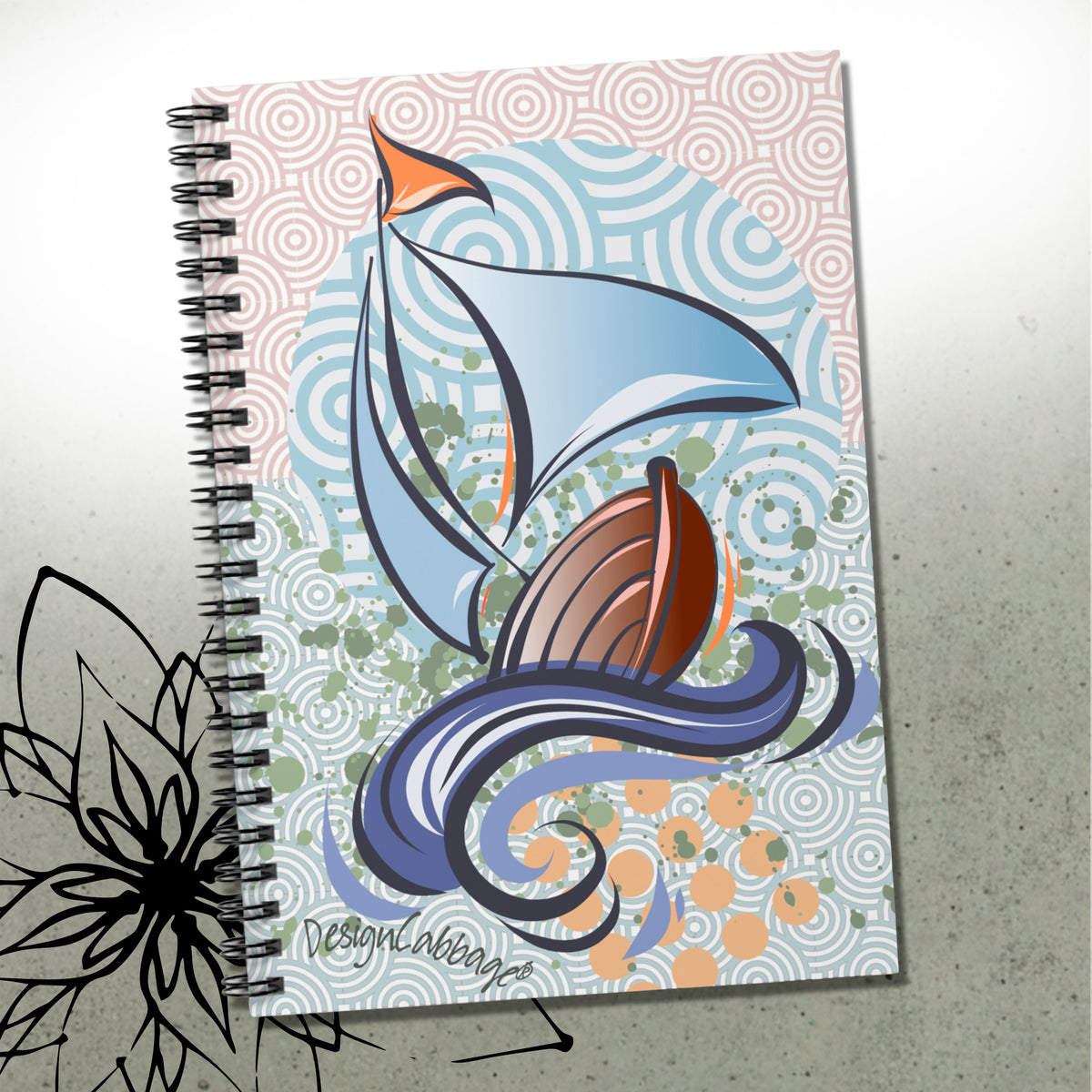 A spiral-bound notebook featuring a stylized illustration of a sailboat with blue and white sails, a brown hull, and swirling ocean waves on a patterned pastel background. The notebook is photographed on a light gray surface with a black floral graphic overlay in the lower left corner.