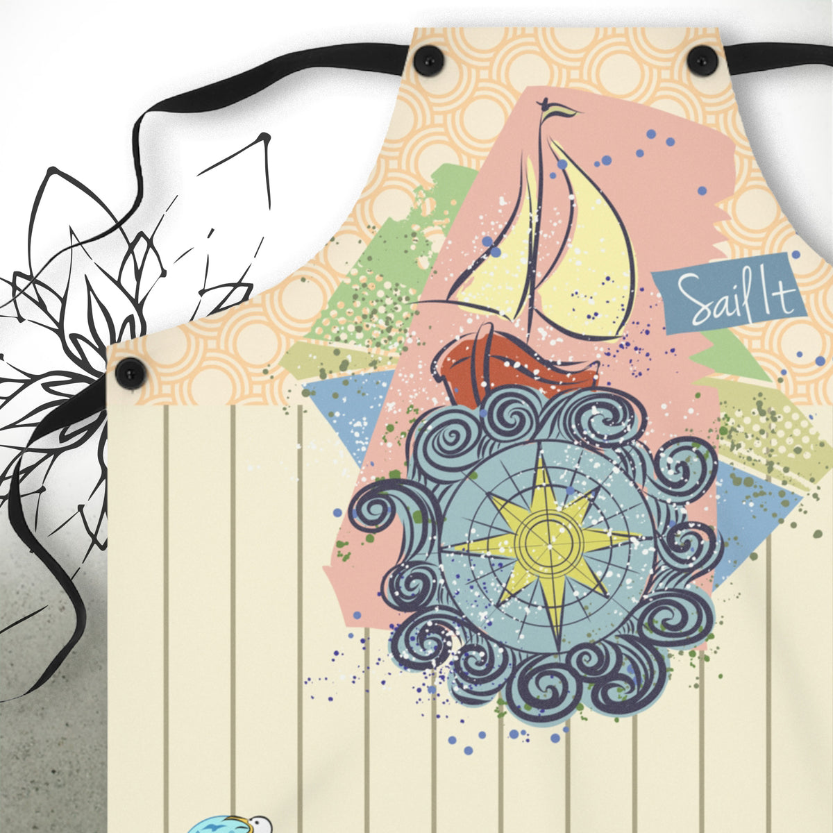 A close-up of a fabric apron with a nautical-themed graphic print. The design features a yellow sailboat, blue ocean waves, a yellow compass rose, and the words 'Sail It' on a pink and beige background.