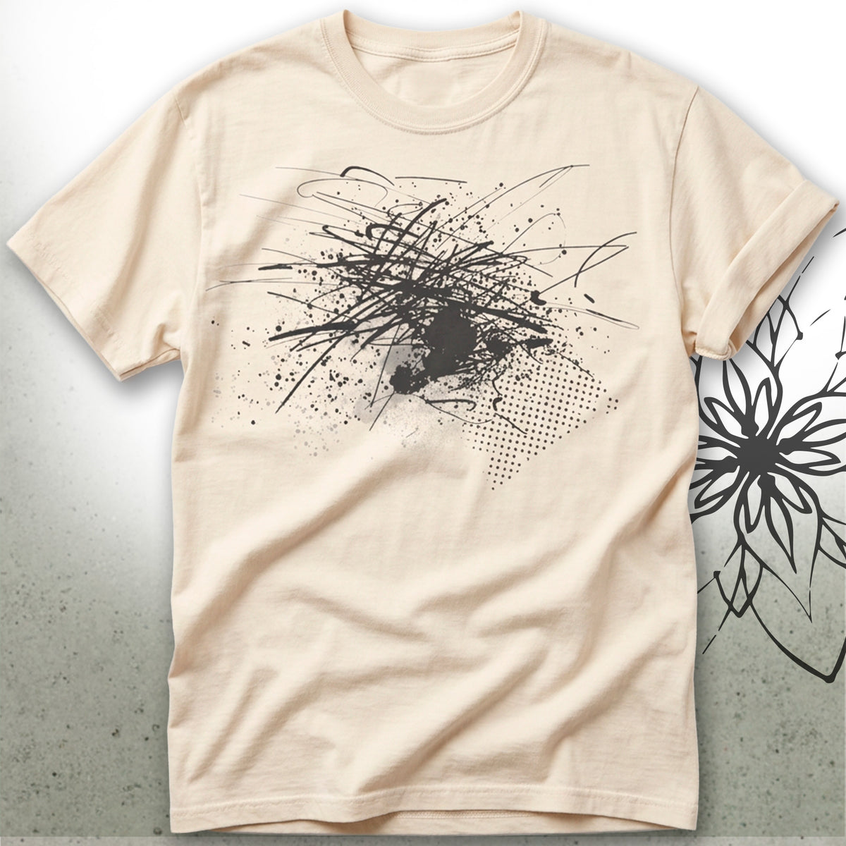 A light beige crewneck t-shirt featuring a black abstract graffiti-style graphic print on the front, displayed against a textured gray background with a black floral illustration on the right.